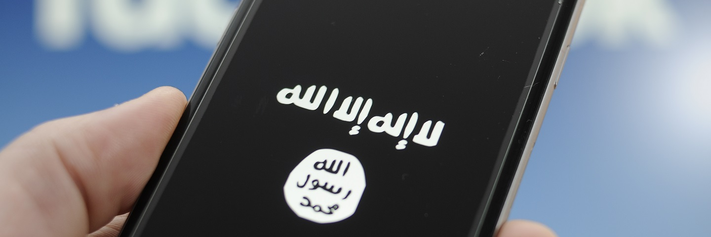The symbol for the terrorist ISIS or Daesh organization is seen on a smartphone. The Facebook logo is blurry in the background.