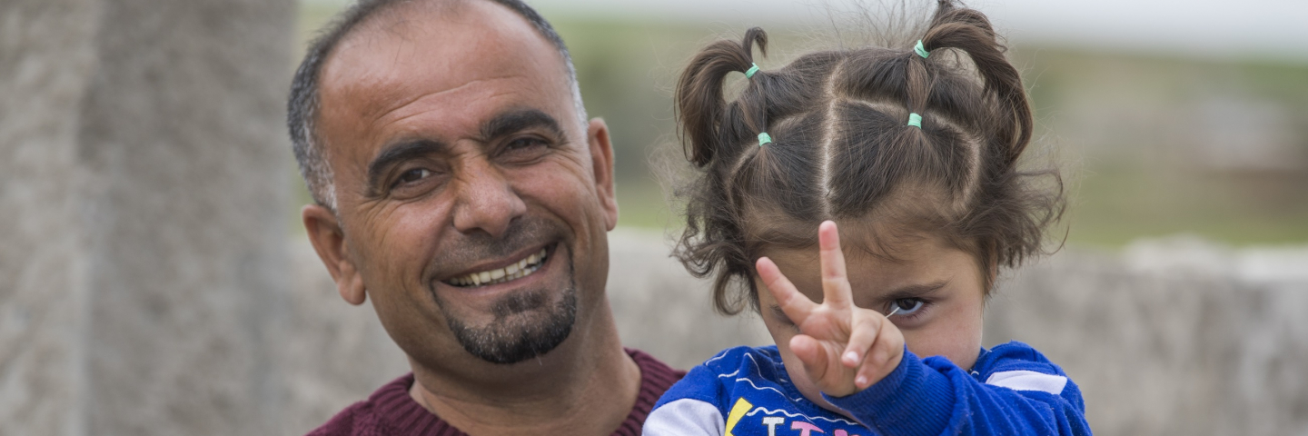 A Syrian child creates the peace sign with her fingers while being held by her father.