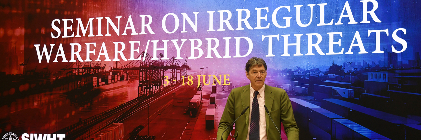 A speaker addresses participants at the George C. Marshall Center during the 2025 Seminar on Irregular Warfare/Hybrid Threats. Behind him, a large digital backdrop displays the course title along with visual elements representing hybrid threats, including cargo ports, drones, and satellites. The event runs from 3–18 June, as shown on screen.