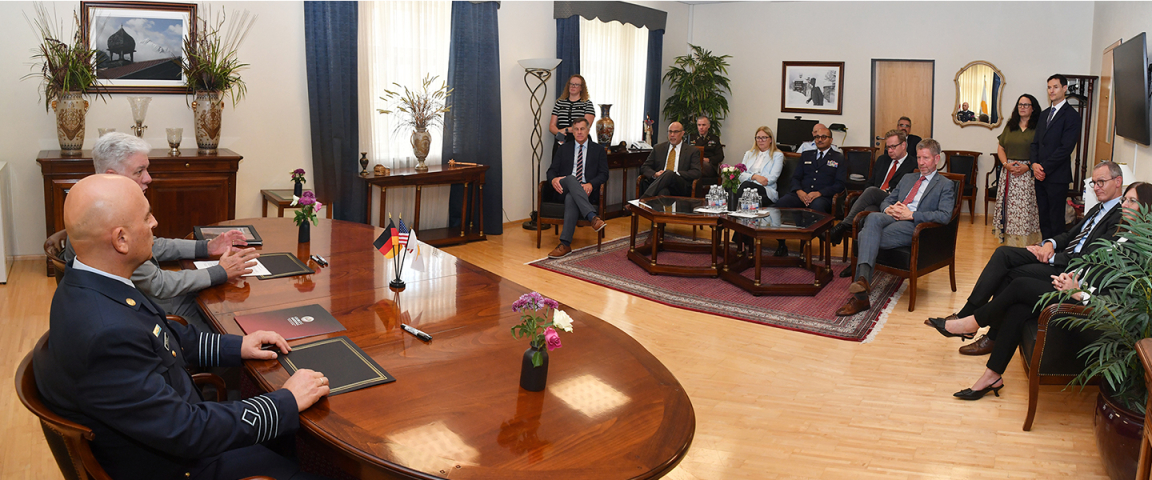 Two men seated at a polished conference table face a room of attendees during a signing ceremony. Small flags from Cyprus, the U.S., and Germany sit on the table. Around the room, about a dozen guests—men and women—observe the exchange, seated in armchairs and standing along the wall.