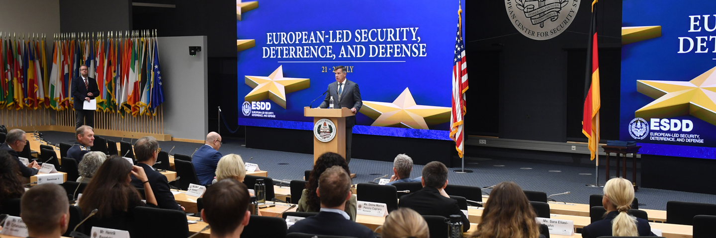 A speaker addresses participants during the Seminar on European-Led Security, Deterrence, and Defense at the George C. Marshall Center. Attendees are seated in a large auditorium, facing a stage flanked by U.S. and German flags and a large screen displaying the seminar title.