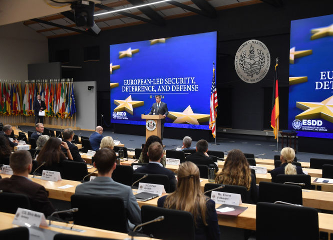 A speaker addresses participants during the Seminar on European-Led Security, Deterrence, and Defense at the George C. Marshall Center. Attendees are seated in a large auditorium, facing a stage flanked by U.S. and German flags and a large screen displaying the seminar title.