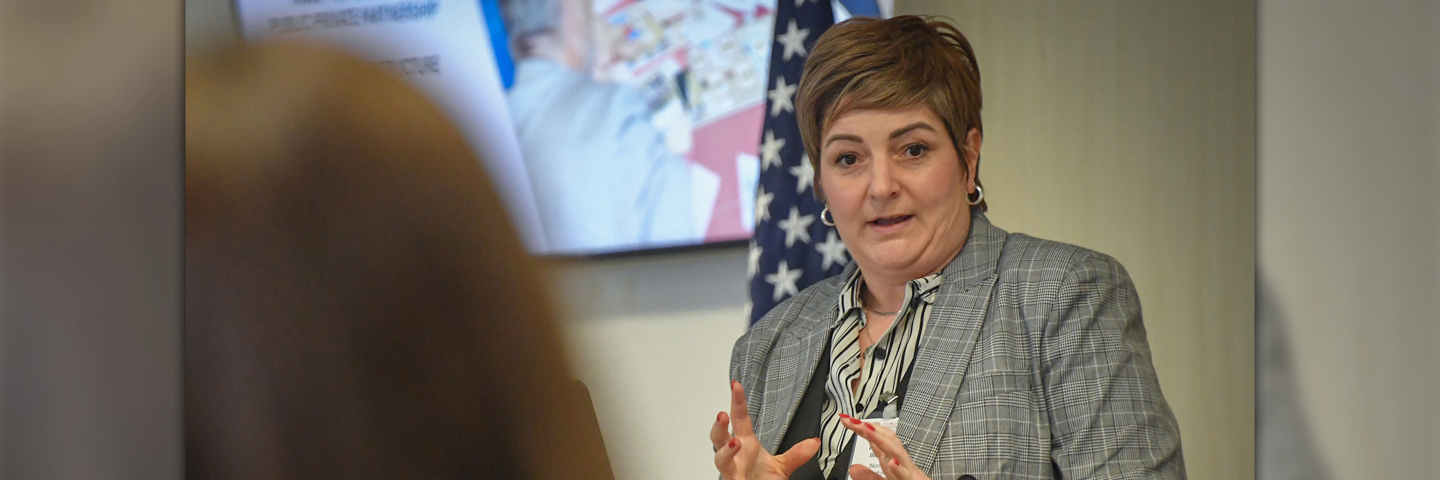 Alumna Scholar Liljana Pecova of North Macedonia speaks during her research presentation at the Marshall Center, gesturing with her hands while standing near an American flag and presentation screen.