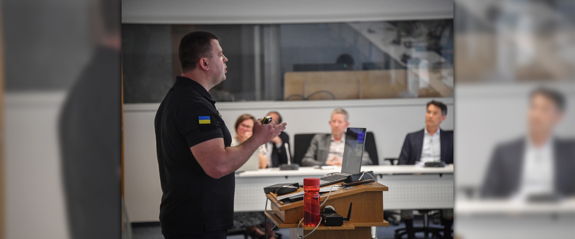 A man in a black shirt with a Ukrainian flag patch presents at a podium in a classroom setting, while several individuals seated at a table listen attentively in the background.
