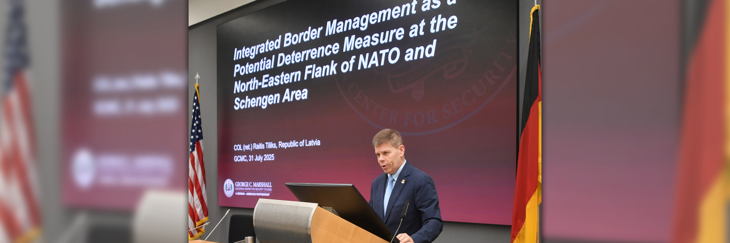 Raitis Tiliks stands at a podium delivering his alumni scholar presentation at the Marshall Center, with a large screen behind him displaying the title of his research on integrated border management and deterrence on NATO’s Northeastern flank