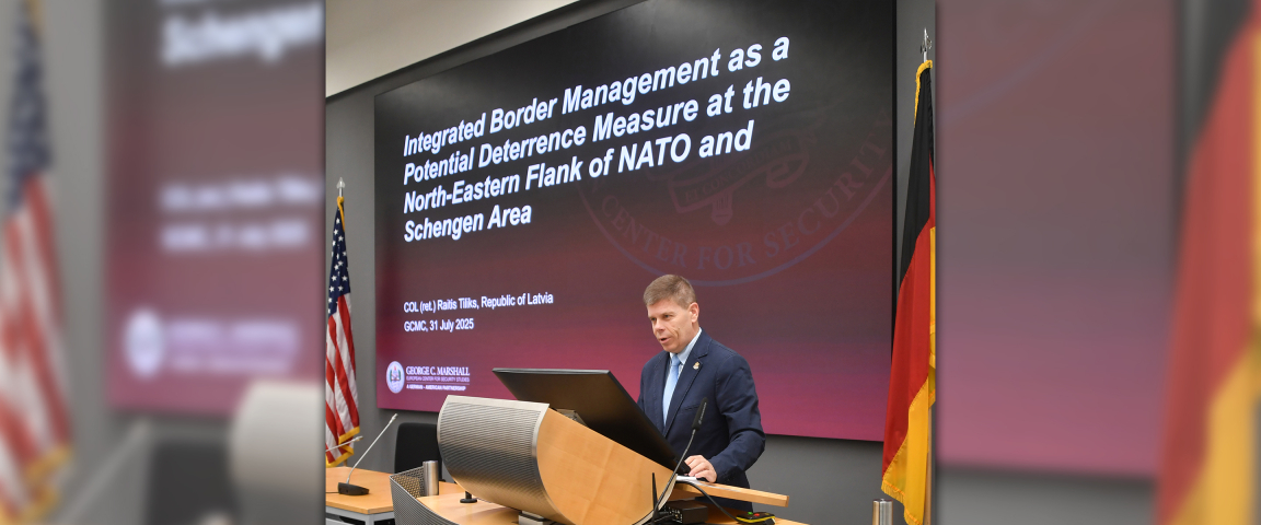 Raitis Tiliks stands at a podium delivering his alumni scholar presentation at the Marshall Center, with a large screen behind him displaying the title of his research on integrated border management and deterrence on NATO’s Northeastern flank