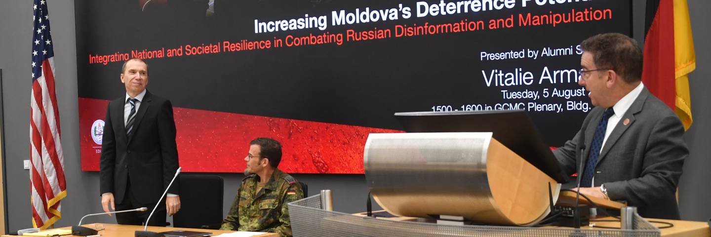 Vitalie Armașu stands at the front of a conference room next to two seated individuals, with a presentation slide behind him titled 'Increasing Moldova's Deterrence Potential.' An American and German flag are visible on either side of the screen.