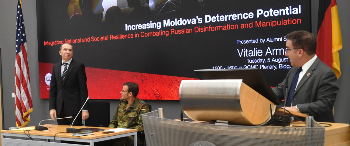 Vitalie Armașu stands at the front of a conference room next to two seated individuals, with a presentation slide behind him titled 'Increasing Moldova's Deterrence Potential.' An American and German flag are visible on either side of the screen.
