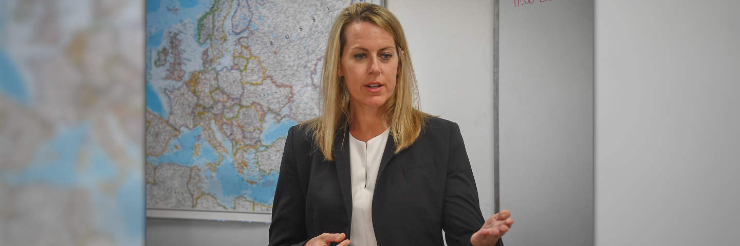 A PLTCE faculty member speaks to participants during a classroom session, gesturing with one hand while standing in front of a map of Europe and a whiteboard.