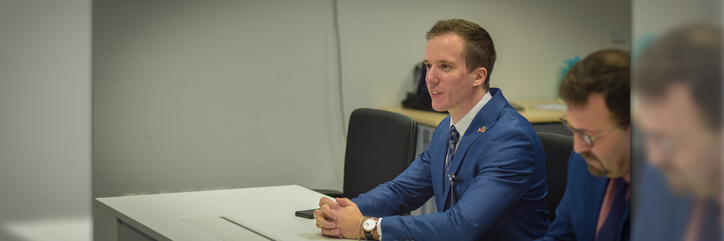 Two men in suits sit at a desk during a Marshall Center seminar. The man in the foreground, wearing a blue suit, speaks while seated, as the second man takes notes.