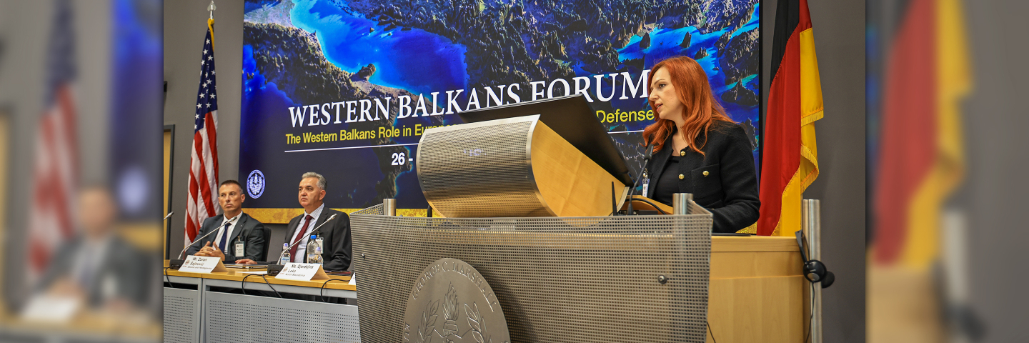 Gjeraqina Leka, assistant professor at the University of Tetova, speaks at the podium during the Western Balkans Forum at the Marshall Center in Garmisch-Partenkirchen, Germany. Behind her, a screen displays the event title alongside the U.S. and German flags. Three panelists are seated at the table to her left.