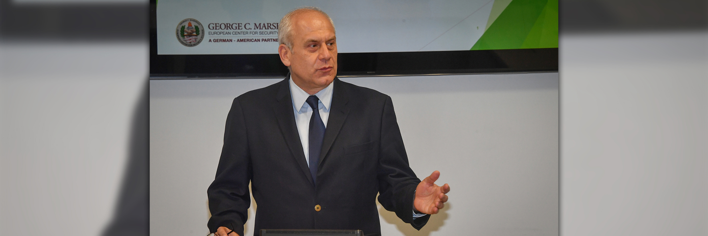 A man stands behind a podium, raising one hand in emphasis as part of a presentation, while in front of a monitor displaying the Marshall Center logo.