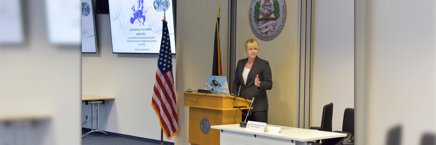 Blonde woman speaks behind a podium with US and German flag behind her.