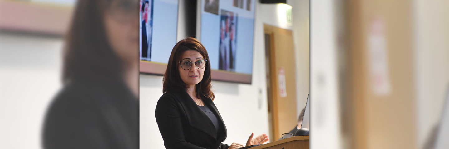 A woman with glasses stands at a podium giving a presentation, with a projection screen visible in the background.