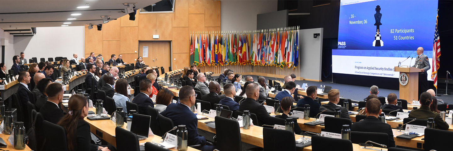 A large group of participants sit in an auditorium facing a stage where a speaker addresses them at a podium. Behind the speaker, a large screen displays the Program on Applied Security Studies dates and participant information, with rows of international flags lining the wall.