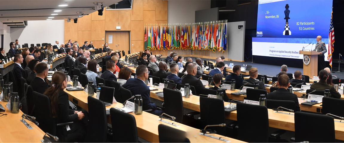 A large group of participants sit in an auditorium facing a stage where a speaker addresses them at a podium. Behind the speaker, a large screen displays the Program on Applied Security Studies dates and participant information, with rows of international flags lining the wall.