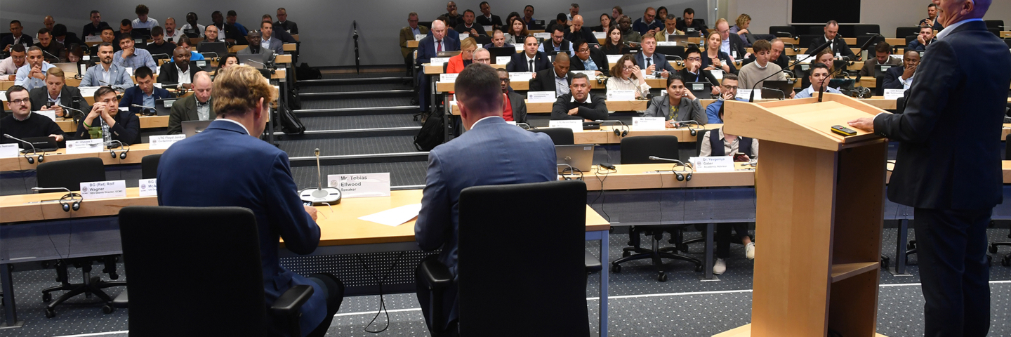 Panelists addresses PASS participants from a podium during the afternoon session at the George C. Marshall Center. The audience of international security professionals sits in rows with laptops and nameplates visible.