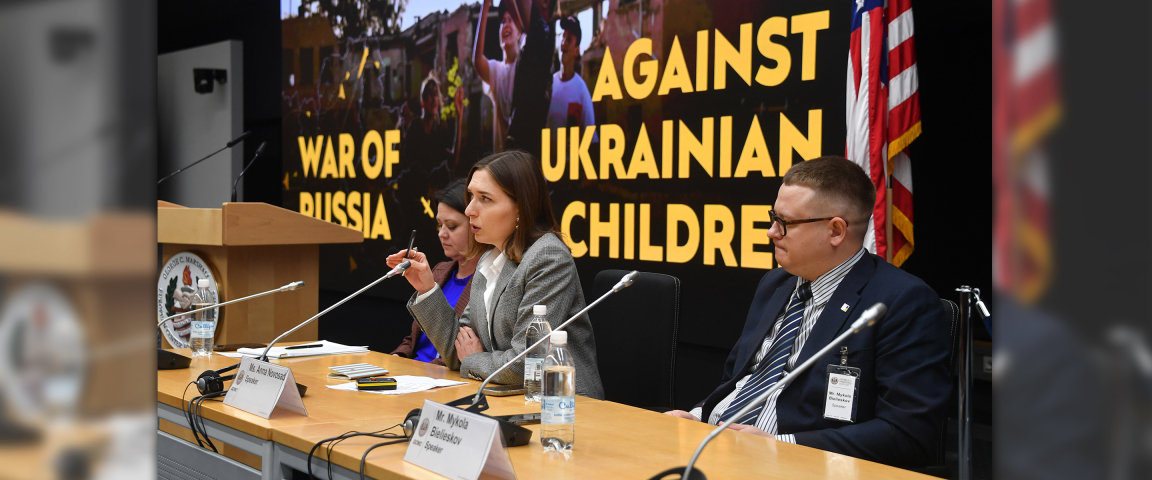 Three speakers sit at a panel table during the Program on Applied Security Studies. A large screen behind them reads “War of Russia Against Ukrainian Children,” and an American flag stands to the right.