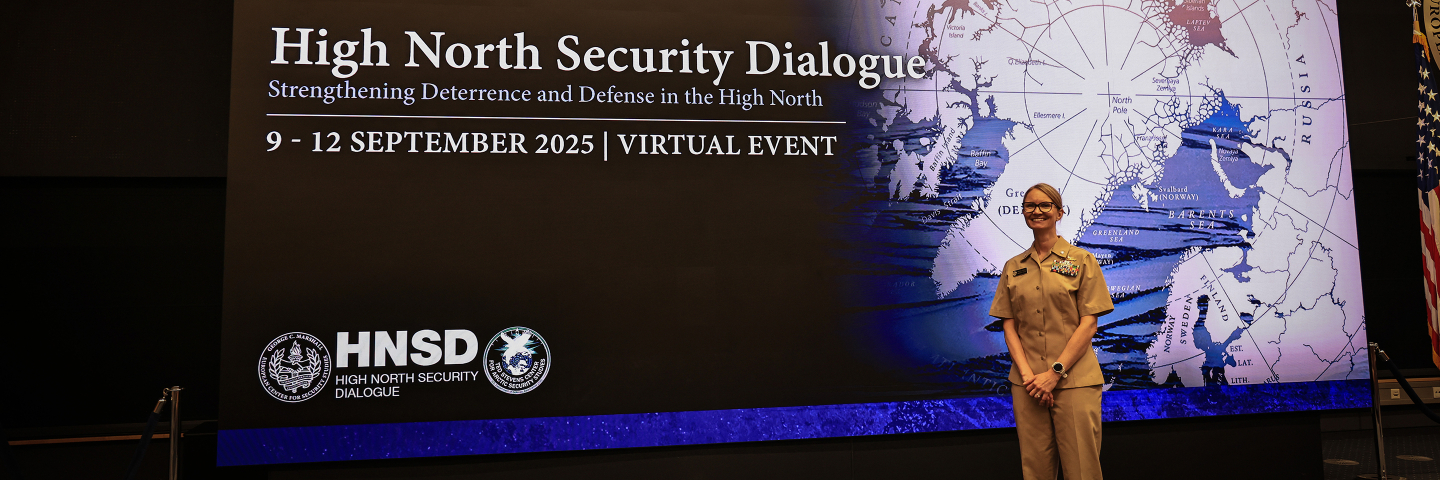 A person in a tan U.S. Navy uniform stands smiling in front of a large screen that reads “High North Security Dialogue” beside a map of the Arctic.
