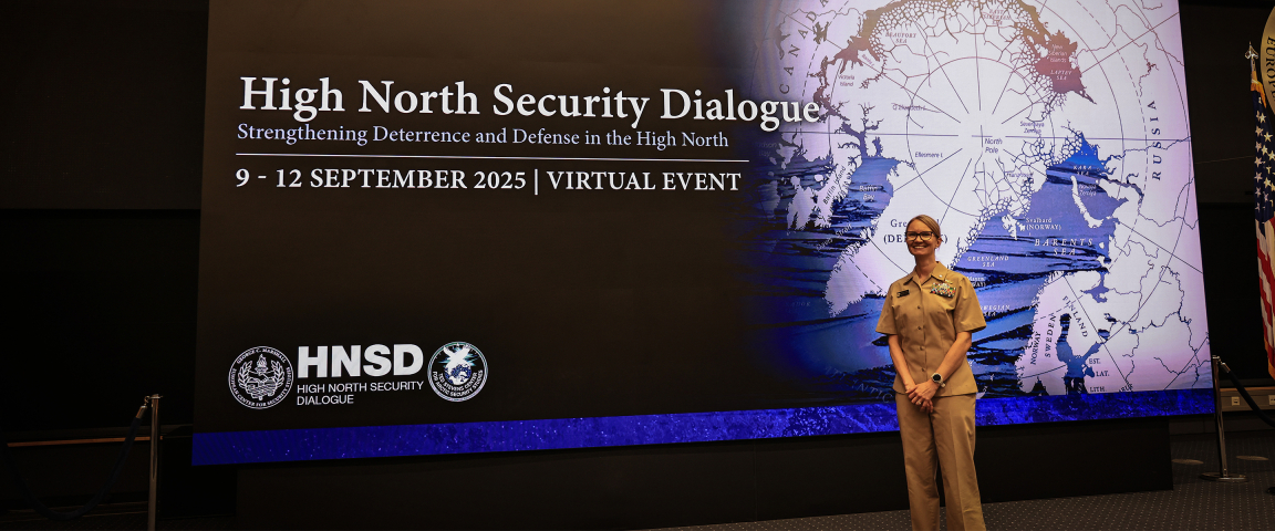 A person in a tan U.S. Navy uniform stands smiling in front of a large screen that reads “High North Security Dialogue” beside a map of the Arctic.