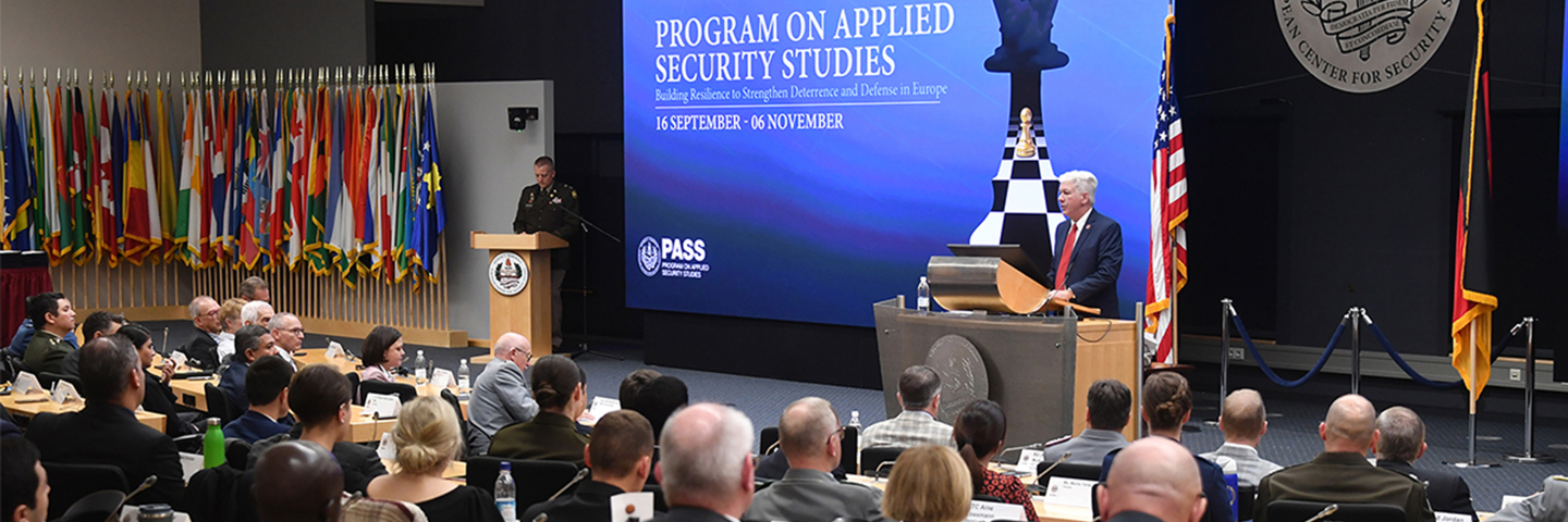 PASS participants sit in a large auditorium as Director Barre Seguin speaks at a podium beside a large course banner and rows of national flags.