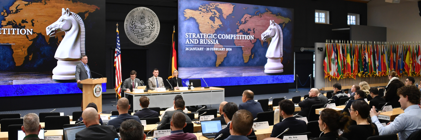 Speaker at a podium addresses participants during the Marshall Center’s “Strategic Competition and Russia” course. A panel sits beside him, with large screens showing a world map and chess knight graphic, and international flags displayed along the wall. 