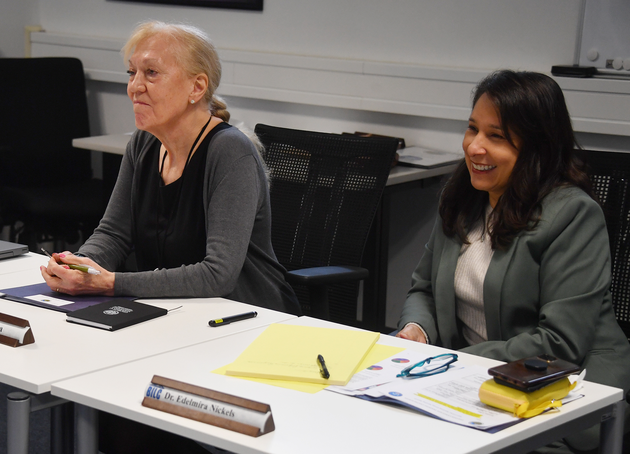 Two women sit at a conference table engaged in discussion. The woman on the left has light hair tied back and wears a dark cardigan, while the woman on the right has long dark hair and is smiling, wearing a gray blazer. Both have papers, pens, and folders in front of them, and a nameplate for "Dr. Edelmira Nickels" is visible on the table. The setting appears to be a professional meeting or seminar in an office or classroom environment.