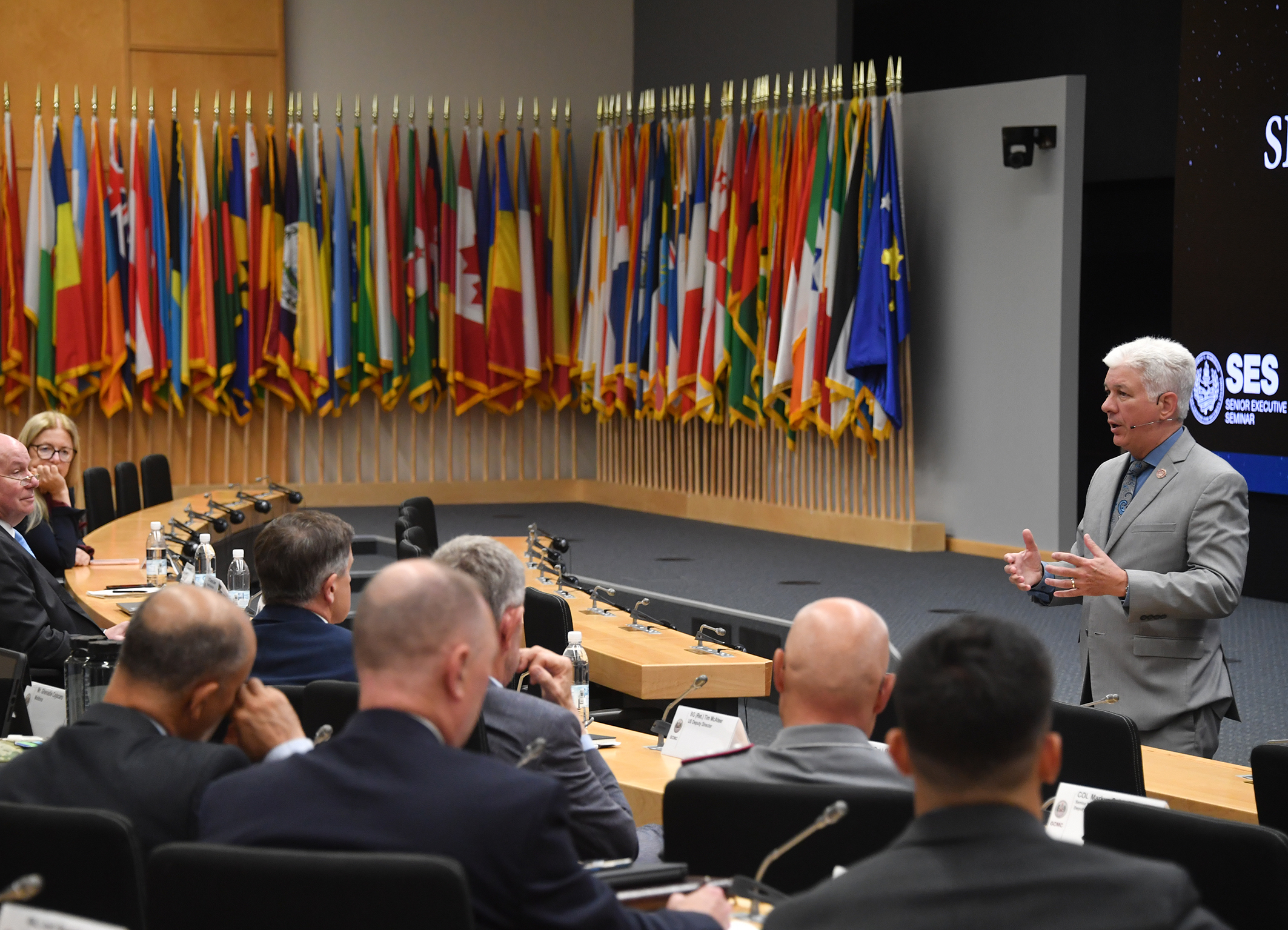 A speaker addresses participants during a session of the Senior Executive Seminar at the George C. Marshall Center. He stands in front of a display featuring the SES logo and speaks to a seated audience. A large display of international flags lines the back of the room, representing the diverse participation in the seminar.