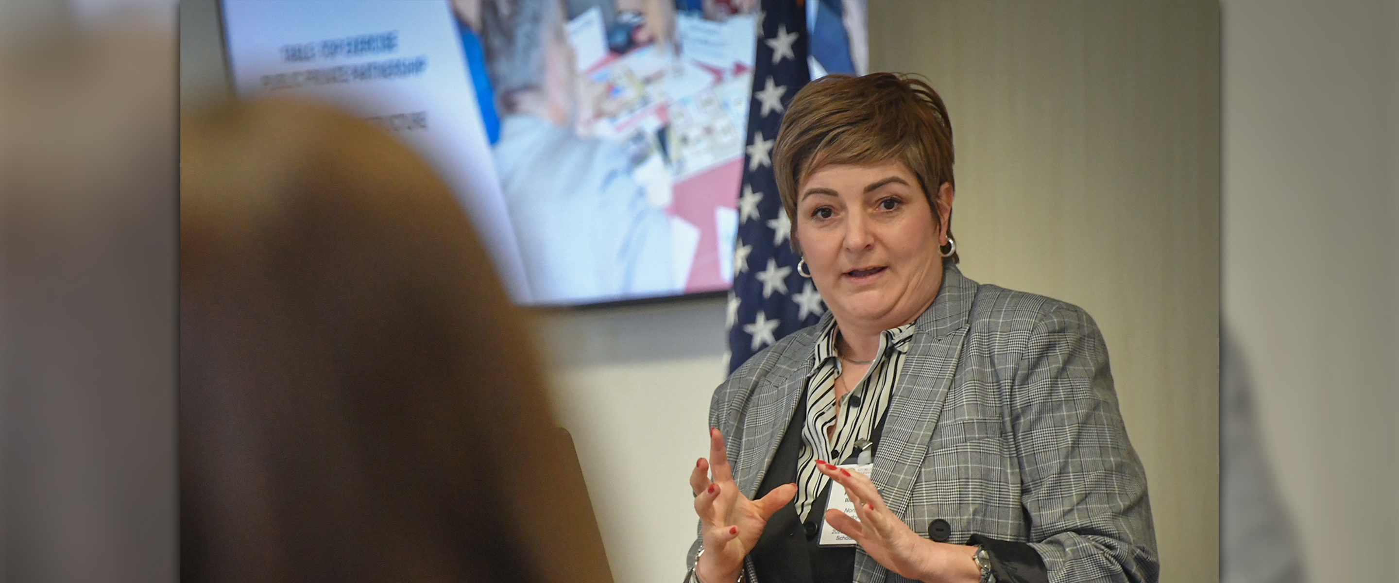 Alumna Scholar Liljana Pecova of North Macedonia speaks during her research presentation at the Marshall Center, gesturing with her hands while standing near an American flag and presentation screen.