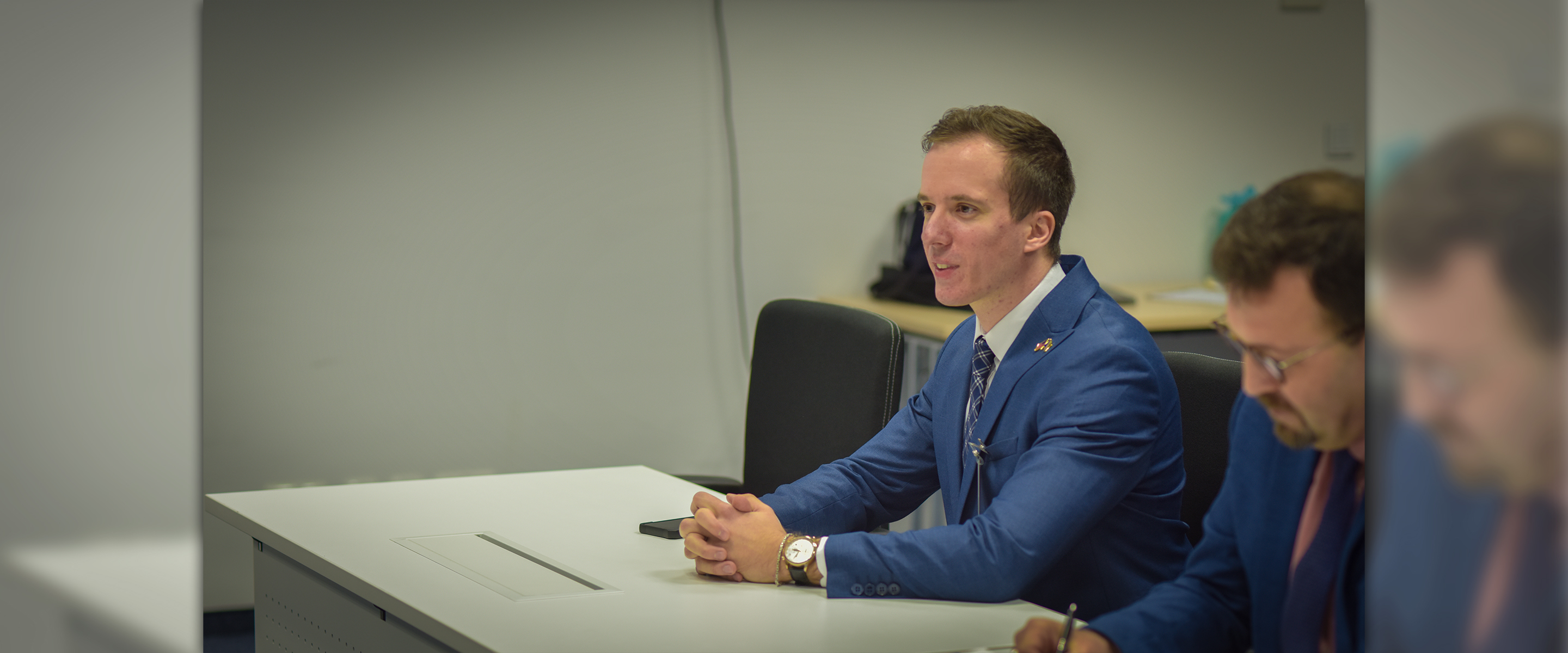 Two men in suits sit at a desk during a Marshall Center seminar. The man in the foreground, wearing a blue suit, speaks while seated, as the second man takes notes.