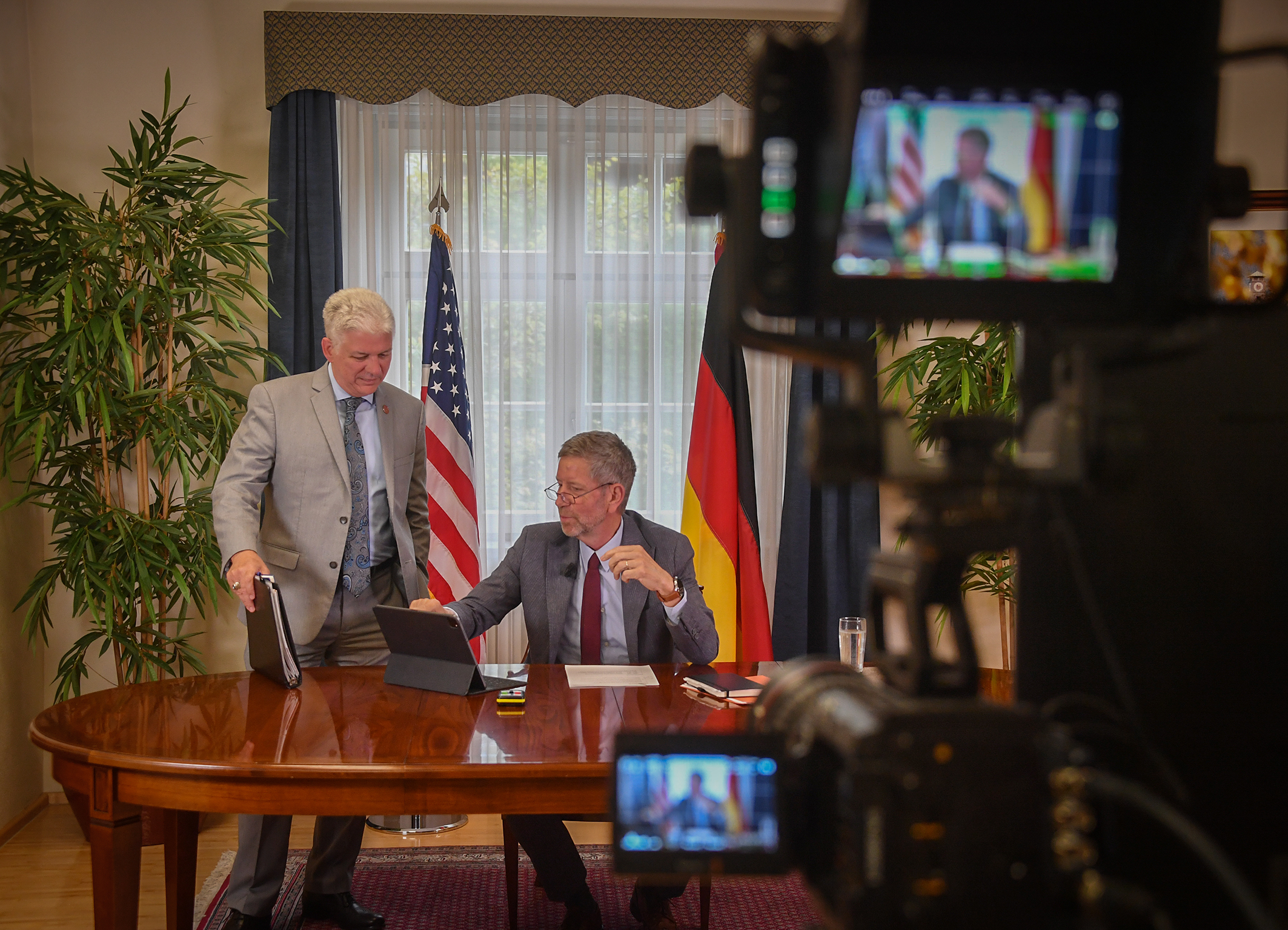 Two men in suits sit and stand at a table with an open tablet, smiling during a meeting. Behind them are U.S. and German flags.