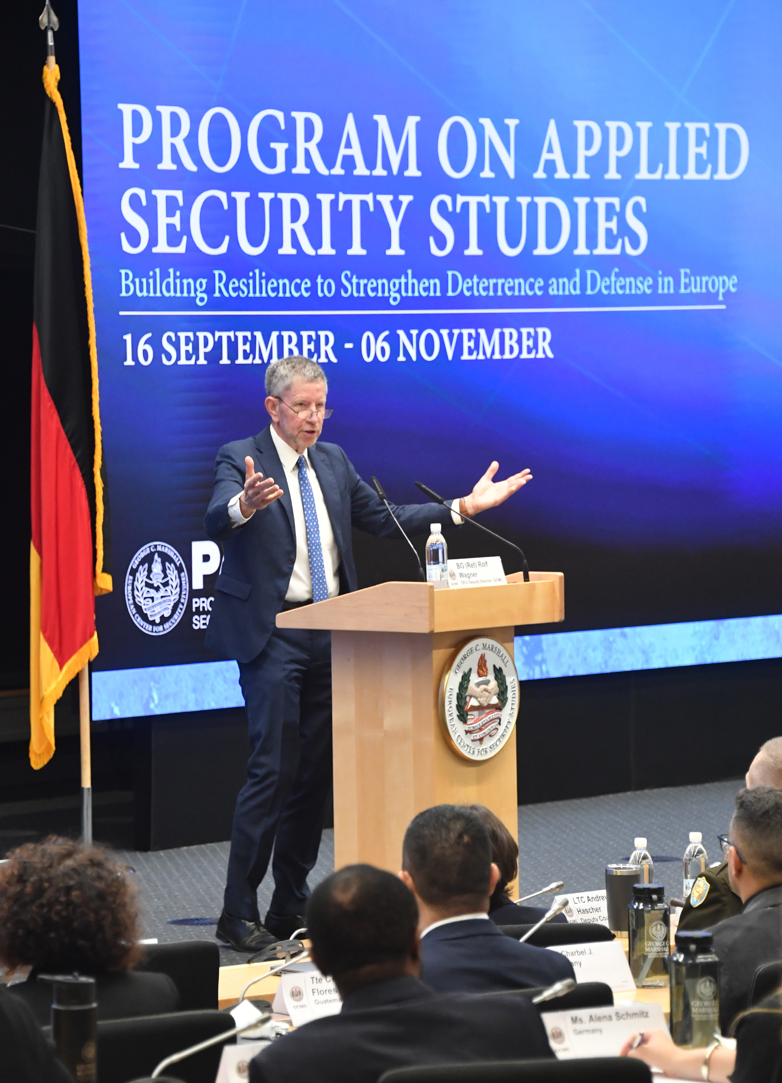 A man stands at a podium addressing participants in an auditorium. Behind him, a large screen displays the title “Program on Applied Security Studies” with the course dates and a chess piece graphic. The U.S. and German flags stand on either side of the stage.