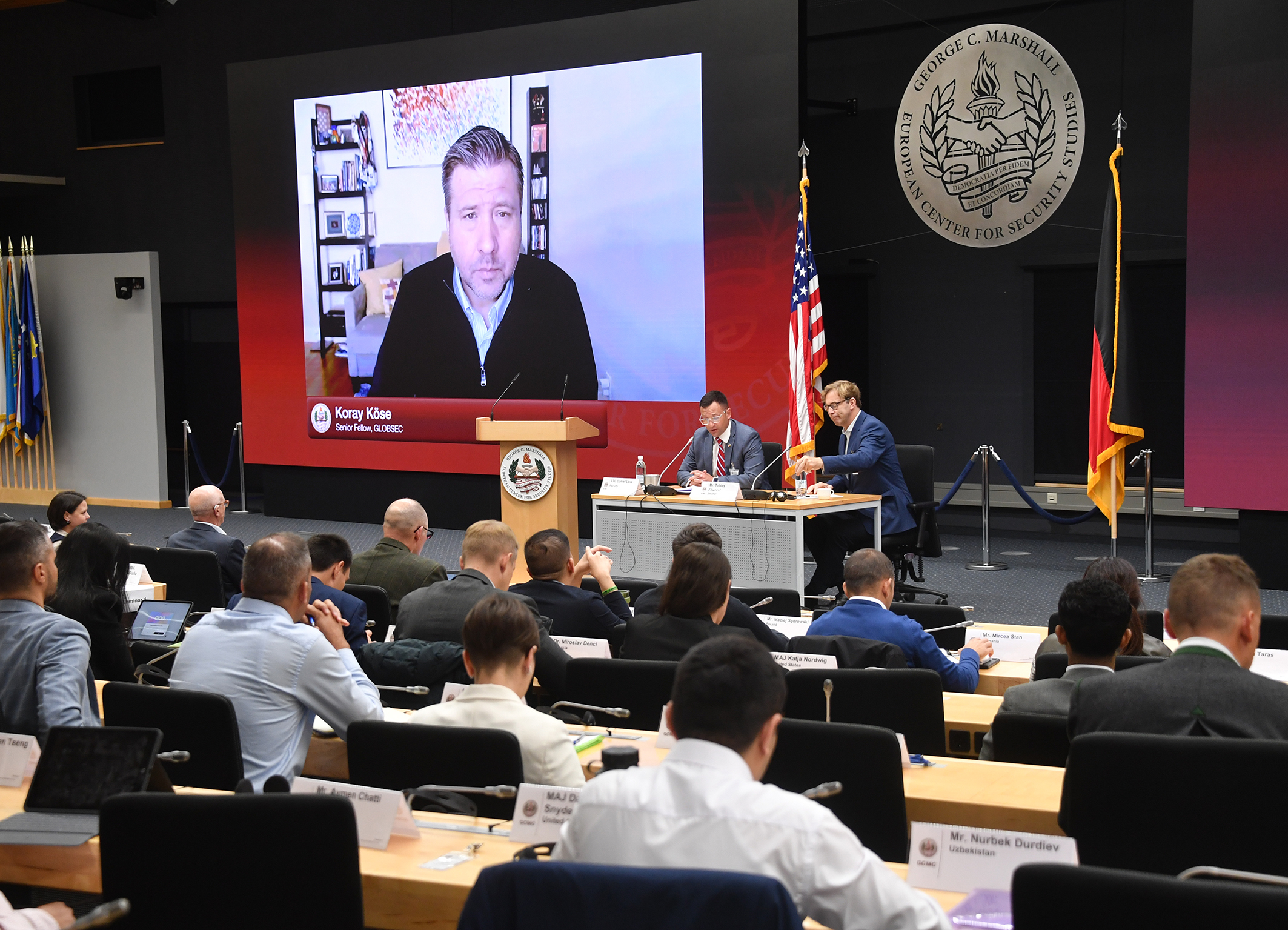 A large screen shows Koray Köse, senior fellow with GLOBSEC, speaking virtually to PASS participants at the George C. Marshall Center. Two panelists are seated at a table in front of the audience, with U.S. and German flags behind them.