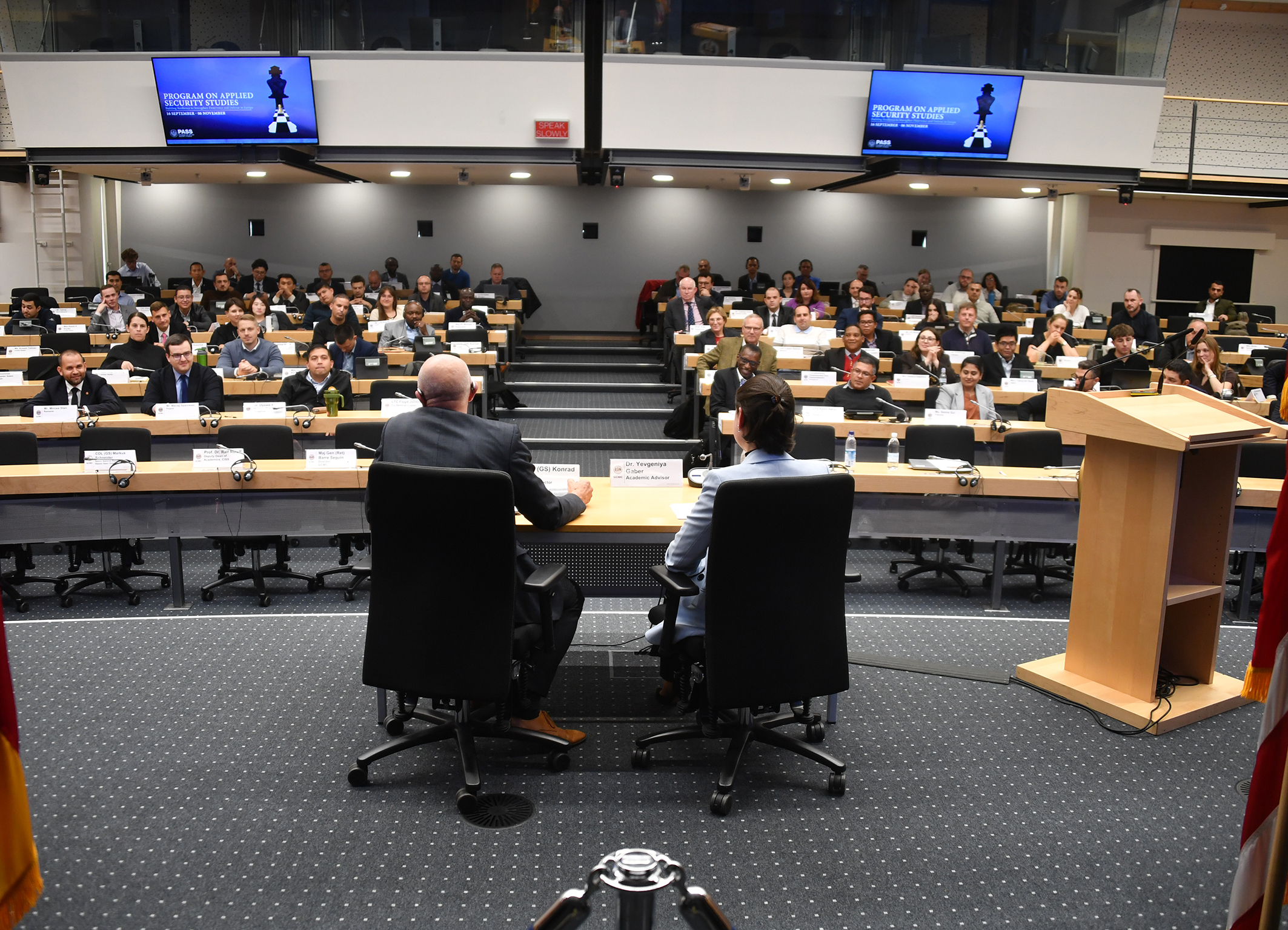 PASS participants sit in a large auditorium facing two speakers seated at a front table during a plenary session.