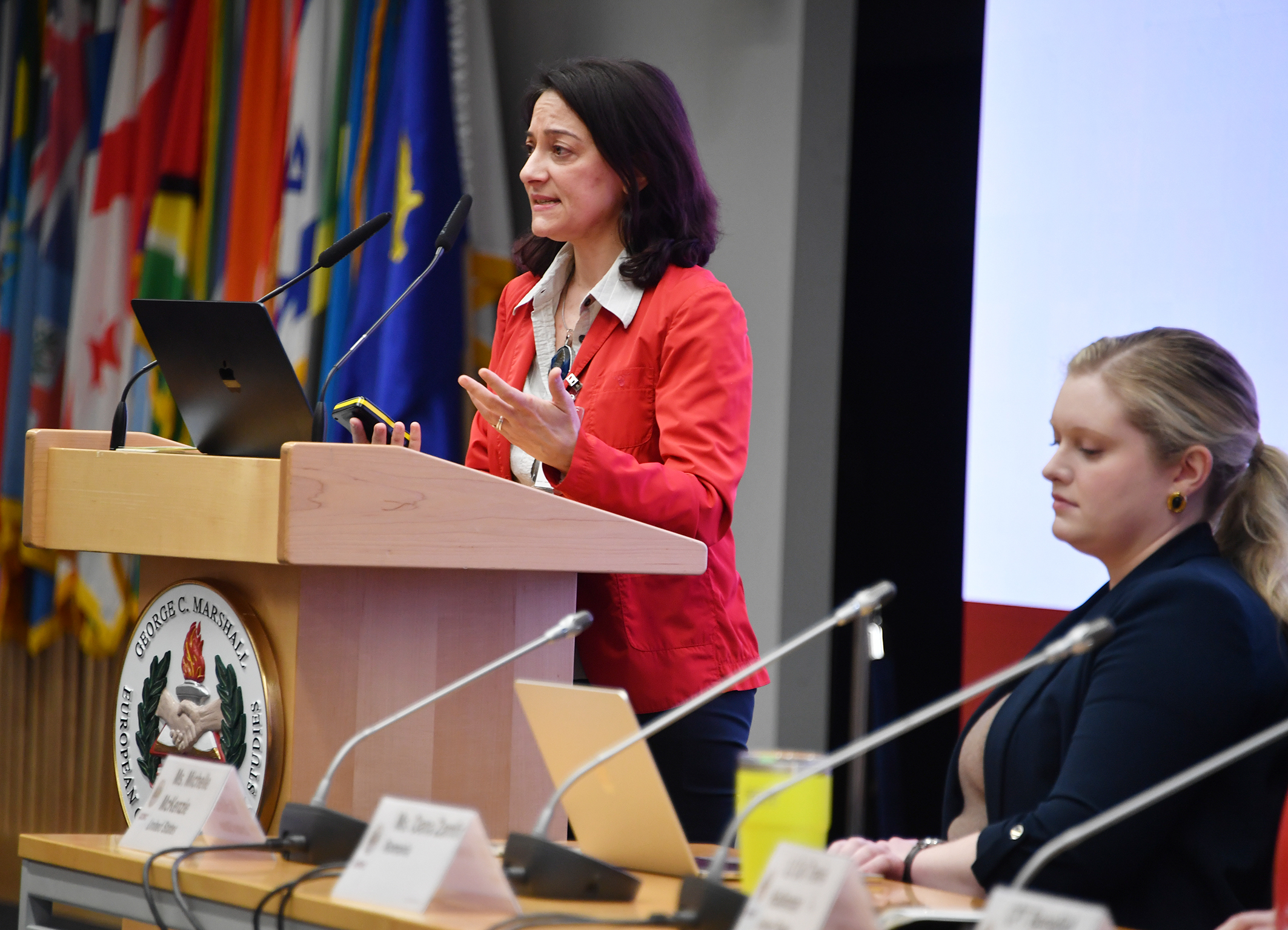 A woman speaks at a podium at the Marshall Center, gesturing as she addresses the audience. Another woman sits beside her at a panel table, with international flags visible in the background.
