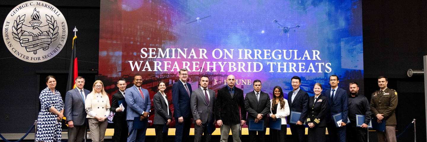 A group of international participants and faculty members pose for a photo on stage in front of a large digital screen displaying the title "Seminar on Irregular Warfare/Hybrid Threats." The backdrop features a blue and red-toned image with drones, satellites, and a cityscape. The George C. Marshall European Center for Security Studies emblem is visible on the wall to the left.