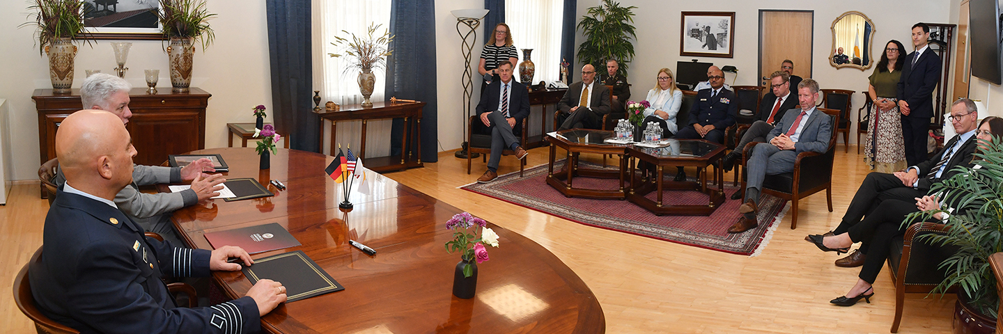 Two men seated at a polished conference table face a room of attendees during a signing ceremony. Small flags from Cyprus, the U.S., and Germany sit on the table. Around the room, about a dozen guests—men and women—observe the exchange, seated in armchairs and standing along the wall.