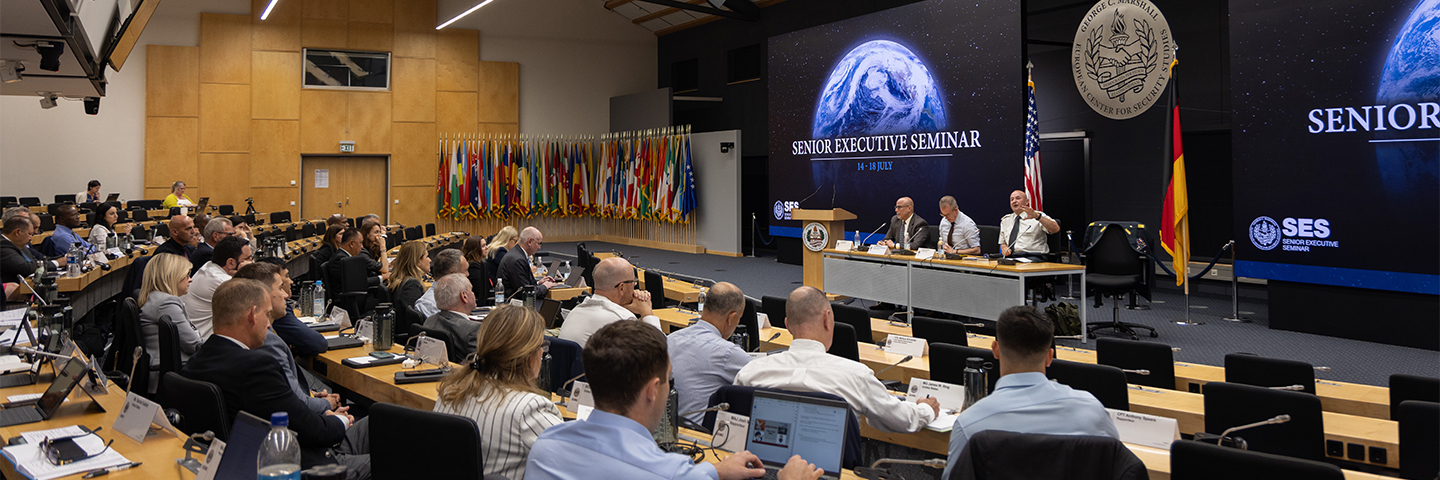 Participants attend a plenary session of the Senior Executive Seminar at the George C. Marshall Center. A panel of speakers sits on stage beneath a large screen displaying the seminar title, “Senior Executive Seminar, 14–18 July.” The auditorium is filled with attendees seated at desks, many using laptops, while a wall of international flags lines the back of the room. U.S. and German flags are displayed near the speakers.