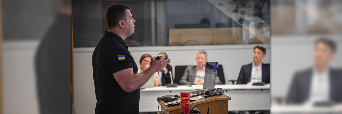 A man in a black shirt with a Ukrainian flag patch presents at a podium in a classroom setting, while several individuals seated at a table listen attentively in the background.