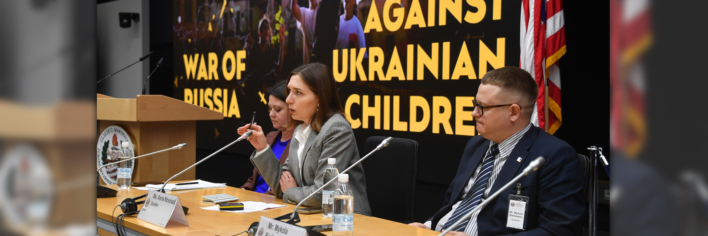 Three speakers sit at a panel table during the Program on Applied Security Studies. A large screen behind them reads “War of Russia Against Ukrainian Children,” and an American flag stands to the right.
