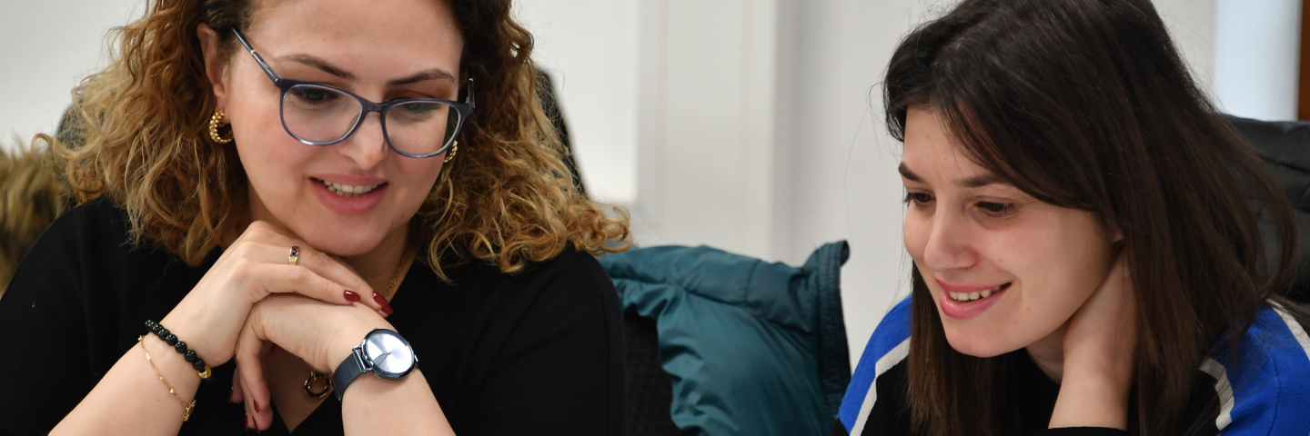 Two women sit together at a table looking at a laptop screen, smiling as they review information during a collaborative discussion.