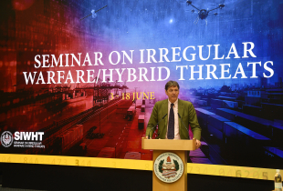 A speaker addresses participants at the George C. Marshall Center during the 2025 Seminar on Irregular Warfare/Hybrid Threats. Behind him, a large digital backdrop displays the course title along with visual elements representing hybrid threats, including cargo ports, drones, and satellites. The event runs from 3–18 June, as shown on screen.