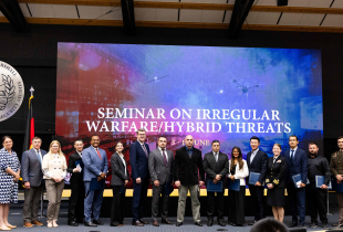 A group of international participants and faculty members pose for a photo on stage in front of a large digital screen displaying the title "Seminar on Irregular Warfare/Hybrid Threats." The backdrop features a blue and red-toned image with drones, satellites, and a cityscape. The George C. Marshall European Center for Security Studies emblem is visible on the wall to the left.