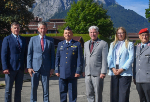 Eight people stand in a line outdoors, dressed in formal business and military attire, posing for a group photo. Behind them are trees, buildings, and a mountain range under a clear blue sky.