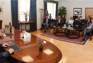 Two men seated at a polished conference table face a room of attendees during a signing ceremony. Small flags from Cyprus, the U.S., and Germany sit on the table. Around the room, about a dozen guests—men and women—observe the exchange, seated in armchairs and standing along the wall.