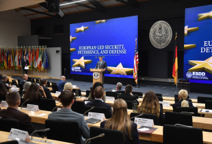 A speaker addresses participants during the Seminar on European-Led Security, Deterrence, and Defense at the George C. Marshall Center. Attendees are seated in a large auditorium, facing a stage flanked by U.S. and German flags and a large screen displaying the seminar title.