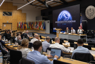 Participants attend a plenary session of the Senior Executive Seminar at the George C. Marshall Center. A panel of speakers sits on stage beneath a large screen displaying the seminar title, “Senior Executive Seminar, 14–18 July.” The auditorium is filled with attendees seated at desks, many using laptops, while a wall of international flags lines the back of the room. U.S. and German flags are displayed near the speakers.