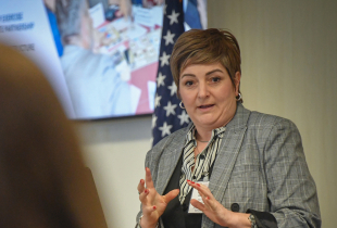 Alumna Scholar Liljana Pecova of North Macedonia speaks during her research presentation at the Marshall Center, gesturing with her hands while standing near an American flag and presentation screen.