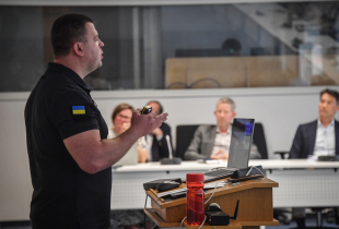 A man in a black shirt with a Ukrainian flag patch presents at a podium in a classroom setting, while several individuals seated at a table listen attentively in the background.
