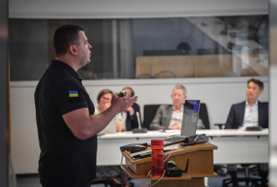 A man in a black shirt with a Ukrainian flag patch presents at a podium in a classroom setting, while several individuals seated at a table listen attentively in the background.